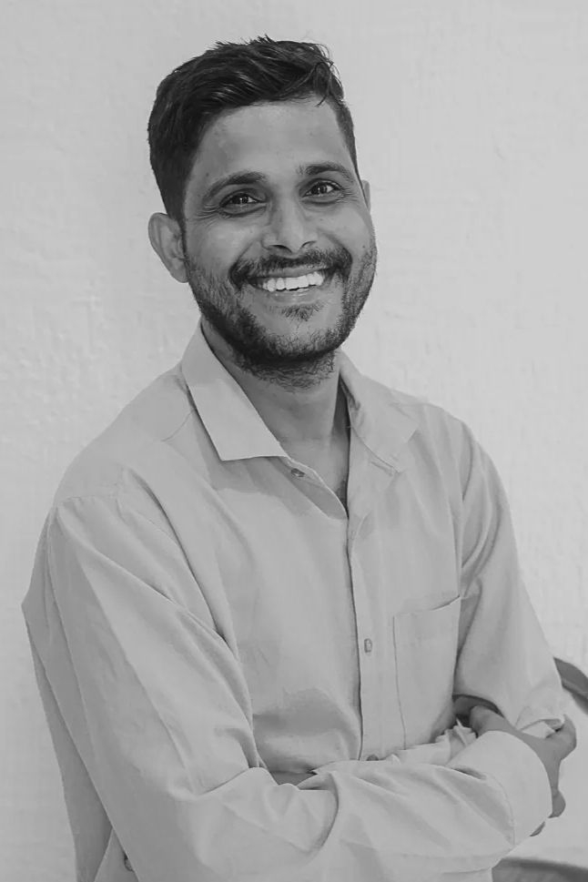 About a smiling man with a beard stands with his arms crossed, wearing a light-colored, button-up shirt against a plain background.