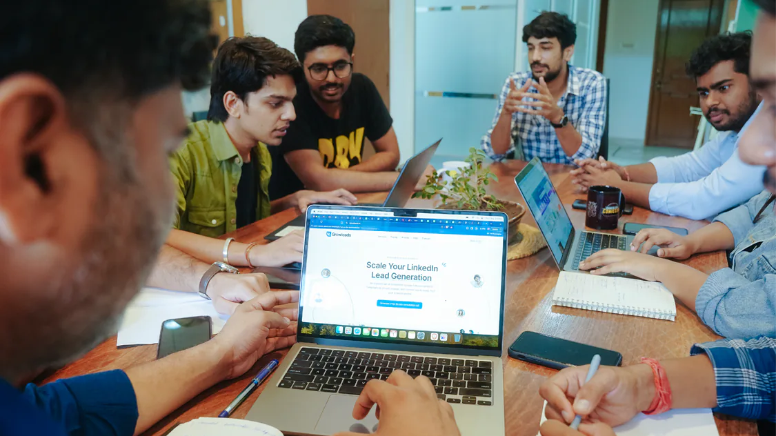 A group of six people sit around a table with laptops and notebooks, engaged in a discussion about 