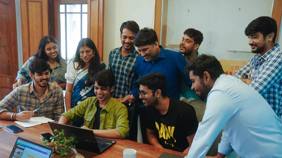 A group of people smiling and looking at a laptop on a wooden table in an indoor setting, discussing about their exciting new project.